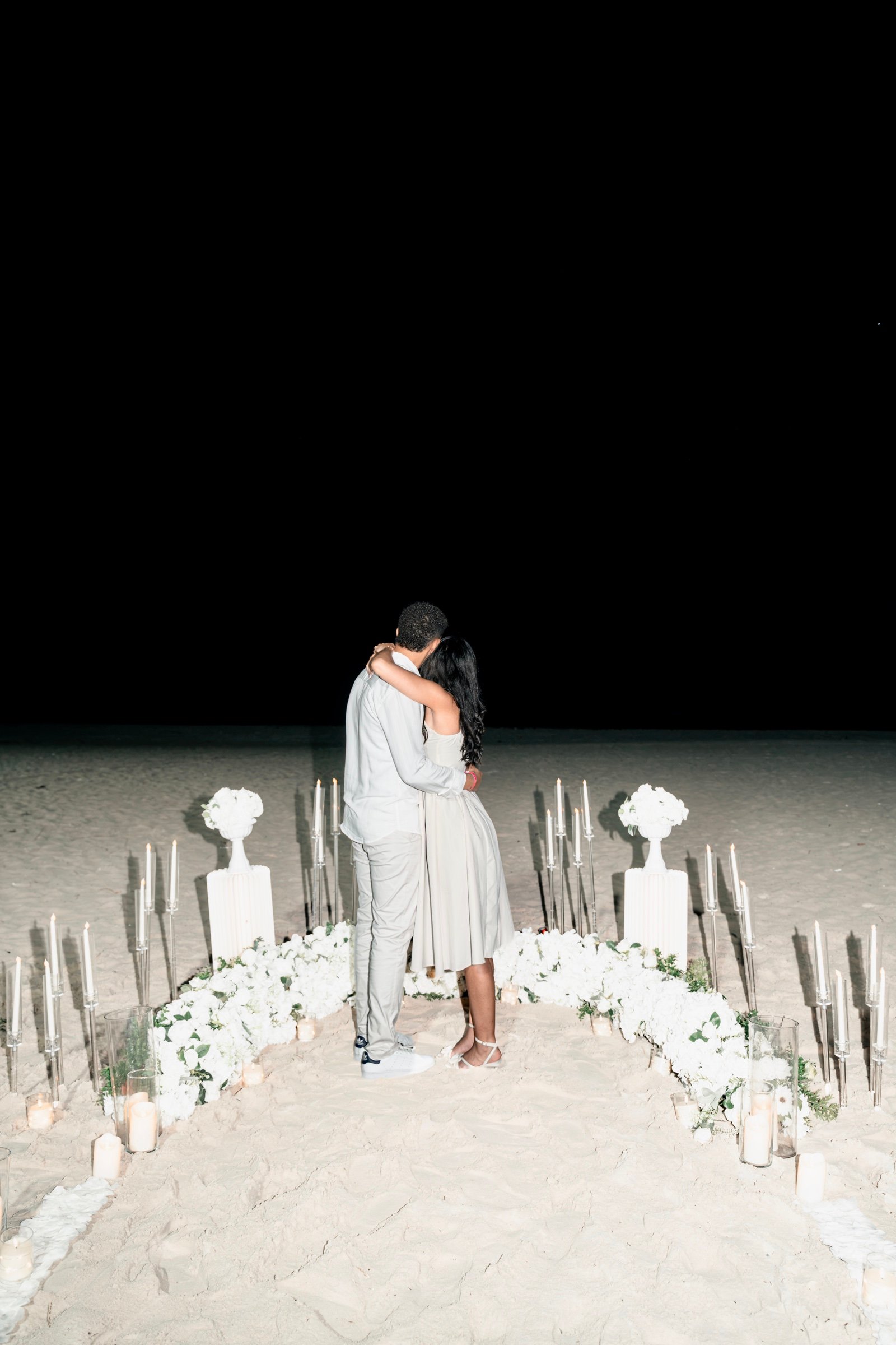 Moises y Sashalee juntos al fondo de un camino de velas y flores blancas.