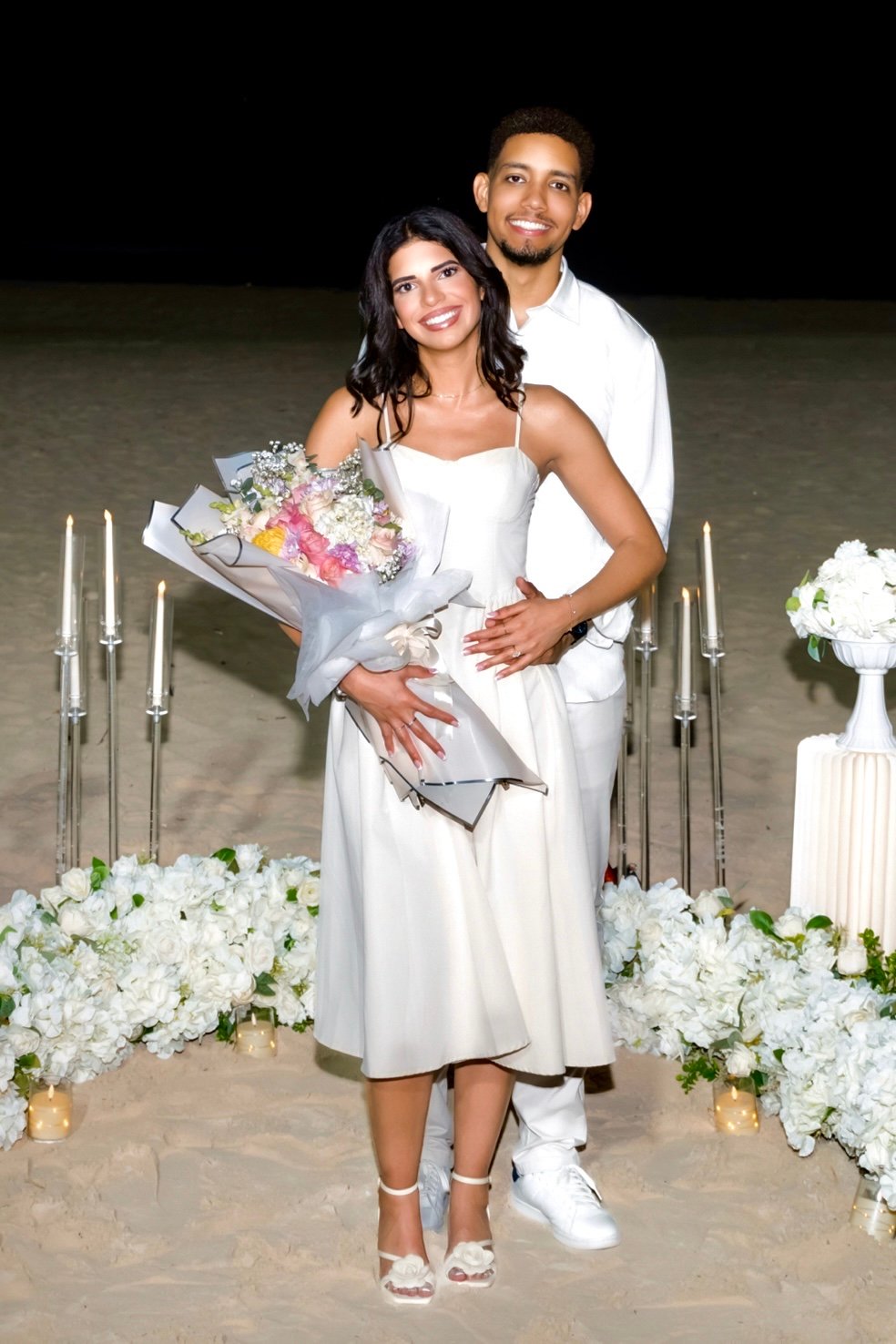 Sashalee sostiene el ramo de flores y Moises la abraza por detrás mientras ambos sonríen.